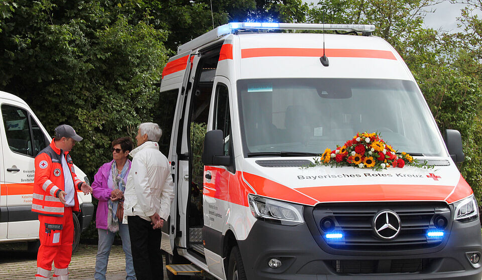 Ein Mann und eine Frau stehen neben einem Einsatzfahrzeug mit Blumenschmuck, ein Mann in Einsatzkleidung erklärt ihnen das Fahrzeug