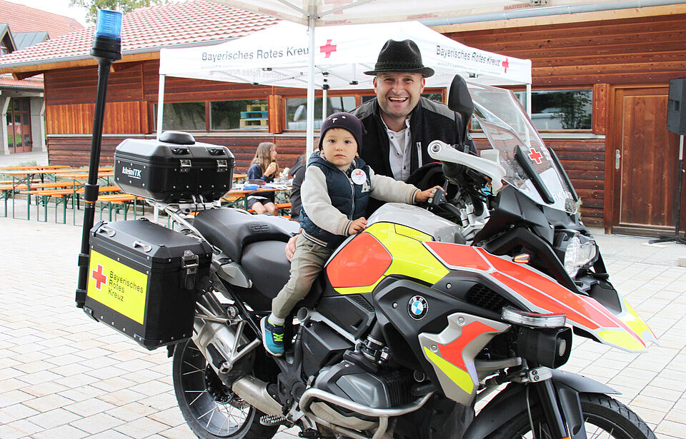 Ein Kind sitzt auf einem Einsatzmotorrad, ein Mann steht dahinter, beide lächeln in die Kamera
