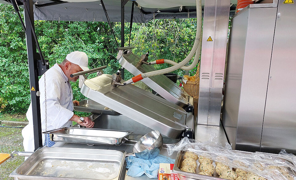 Ein Mann in weißer Kochjacke steht am geöffneten Kessel einer Feldküche