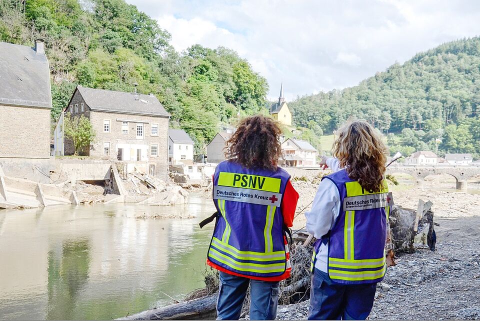Zwei Helferinnen mit Kennzeichnungswesten mit der Aufschrift PSNV stehen 2021 am Ufer der Ahr und blicken auf die Zerstörung durch das Hochwasser